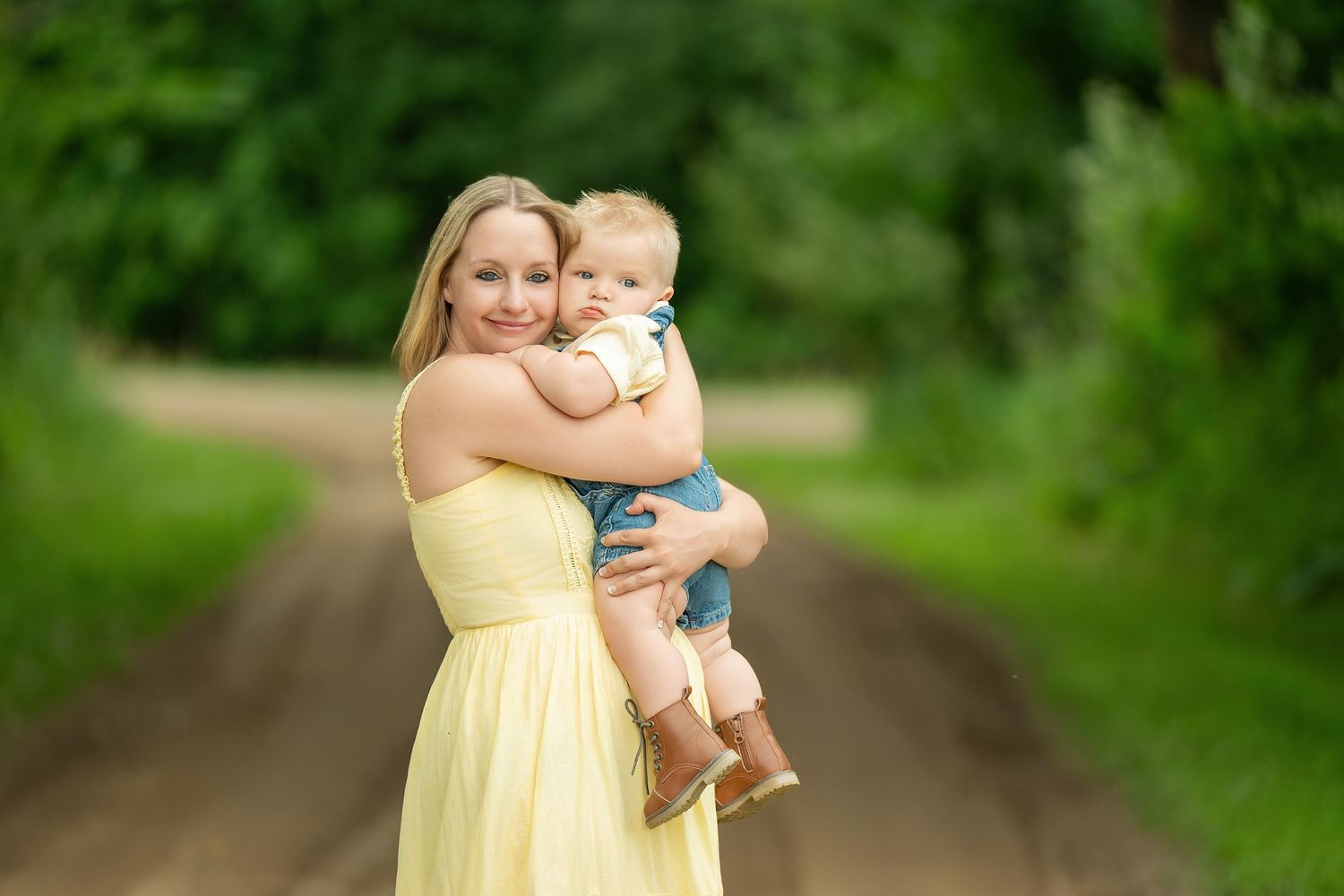 A parent in a yellow sundress holds a baby on a dirt path surrounded by lush greenery. Family Portraits & Milestone Moments in Foreston, MN