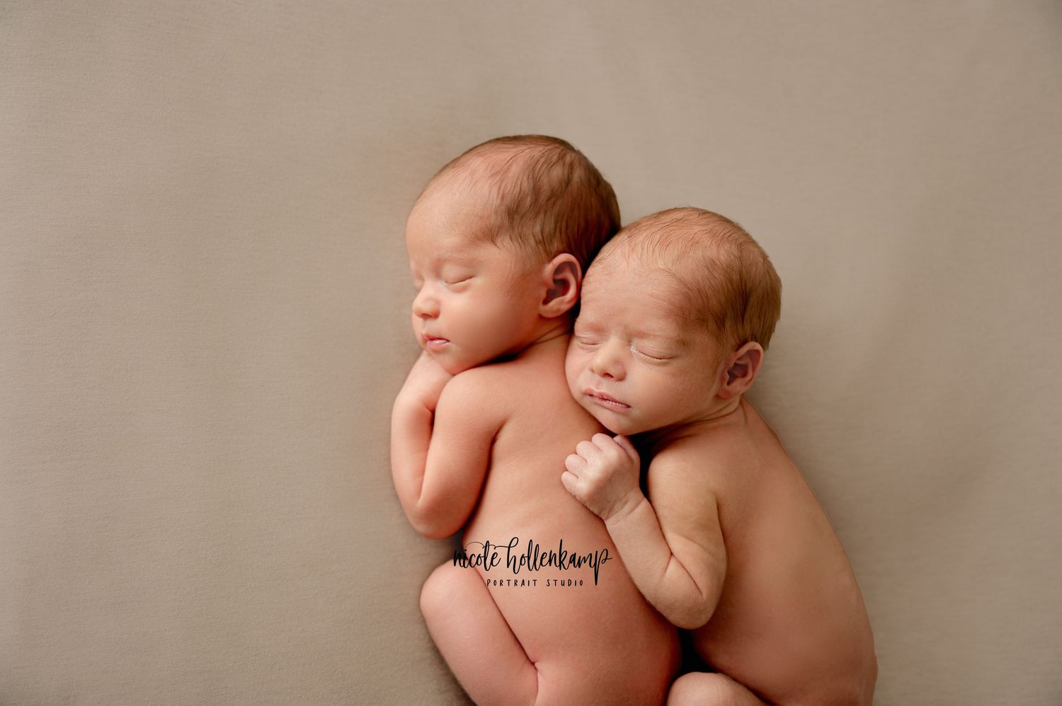 Two sleeping newborn twins posed close together on soft beige backdrop in studio setting.