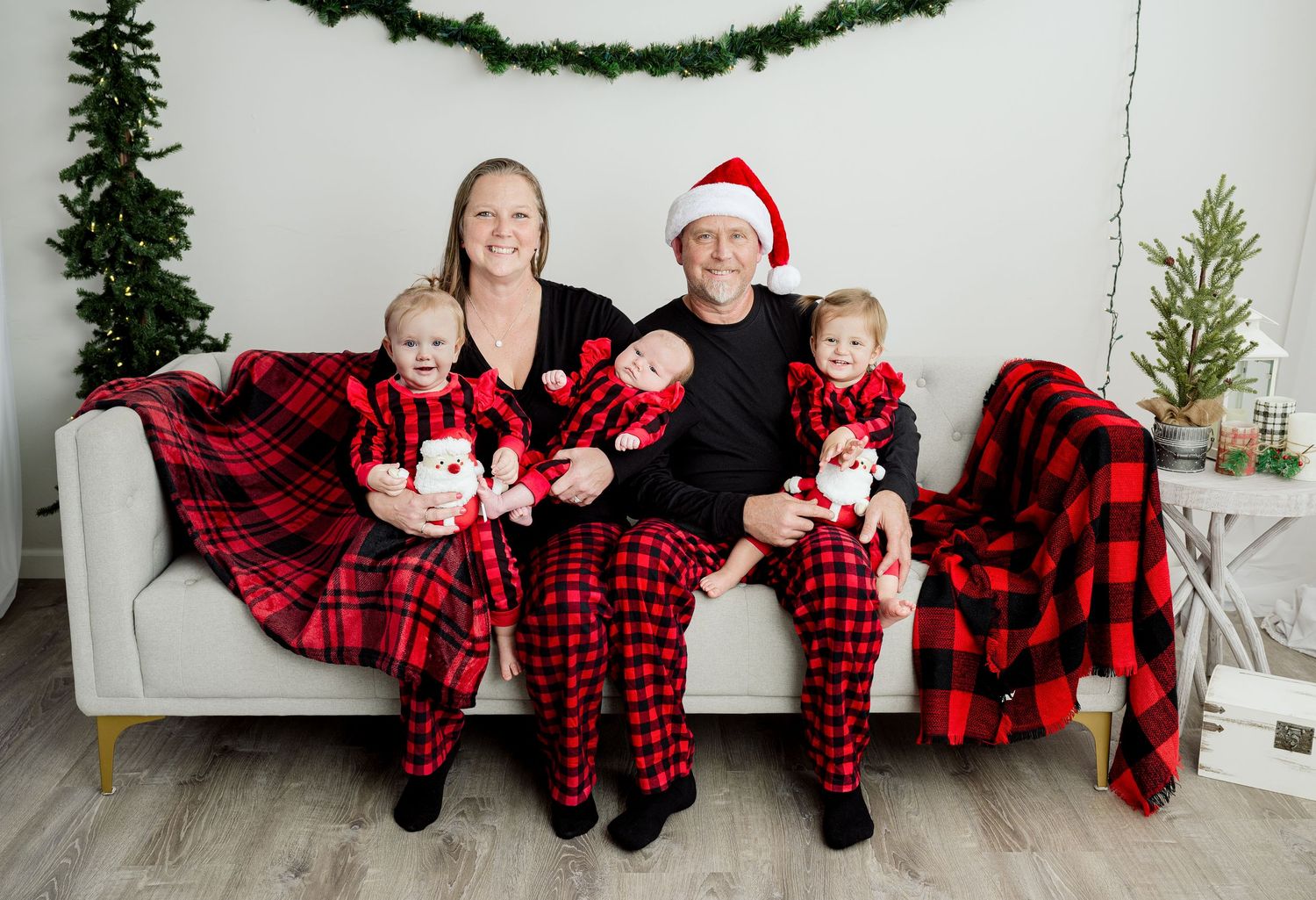 Cozy family Christmas photo on white couch wearing matching buffalo plaid pajamas with holiday decorations and greenery.