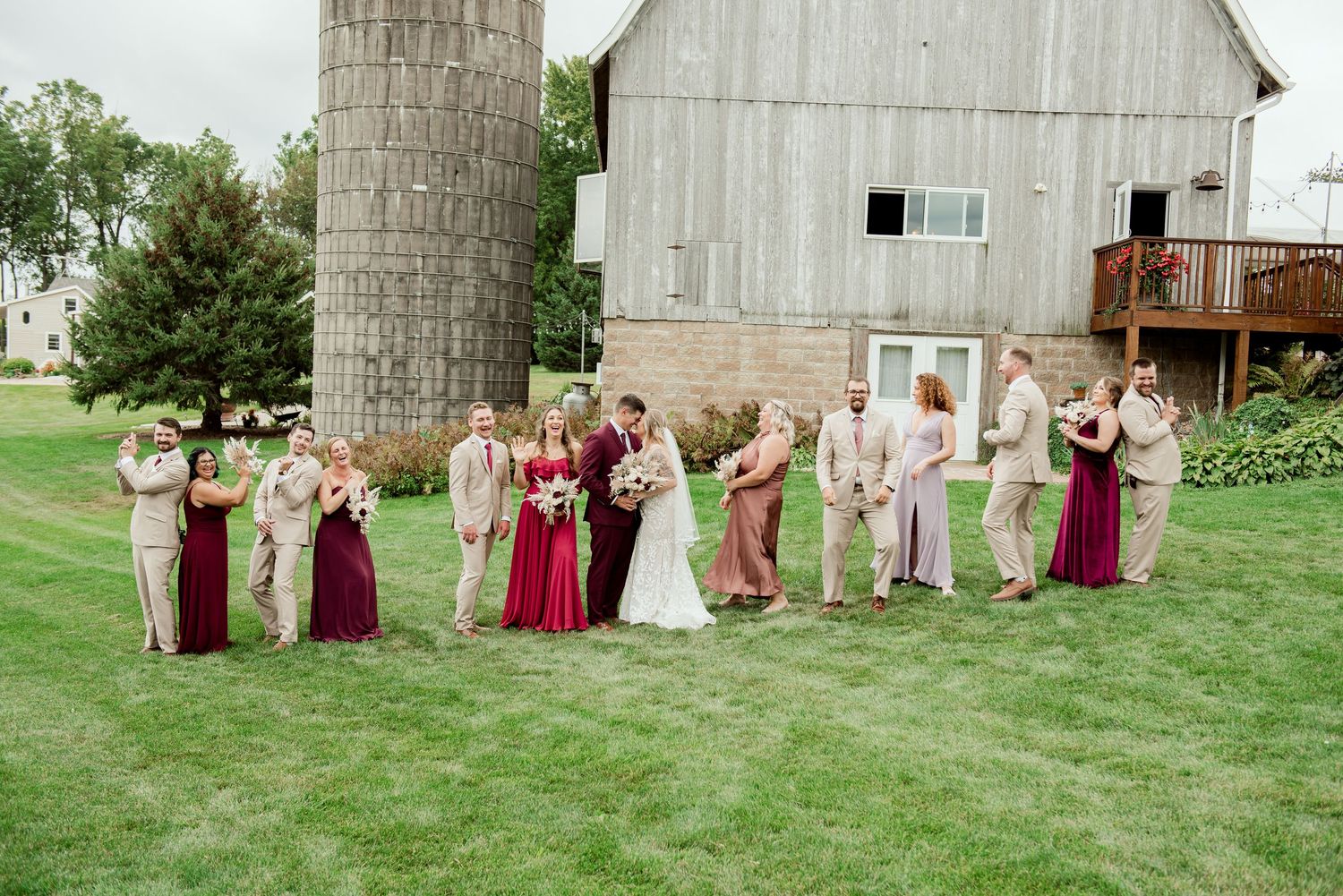 Wedding group poses for casual photos in front of rustic barn and stone silo.