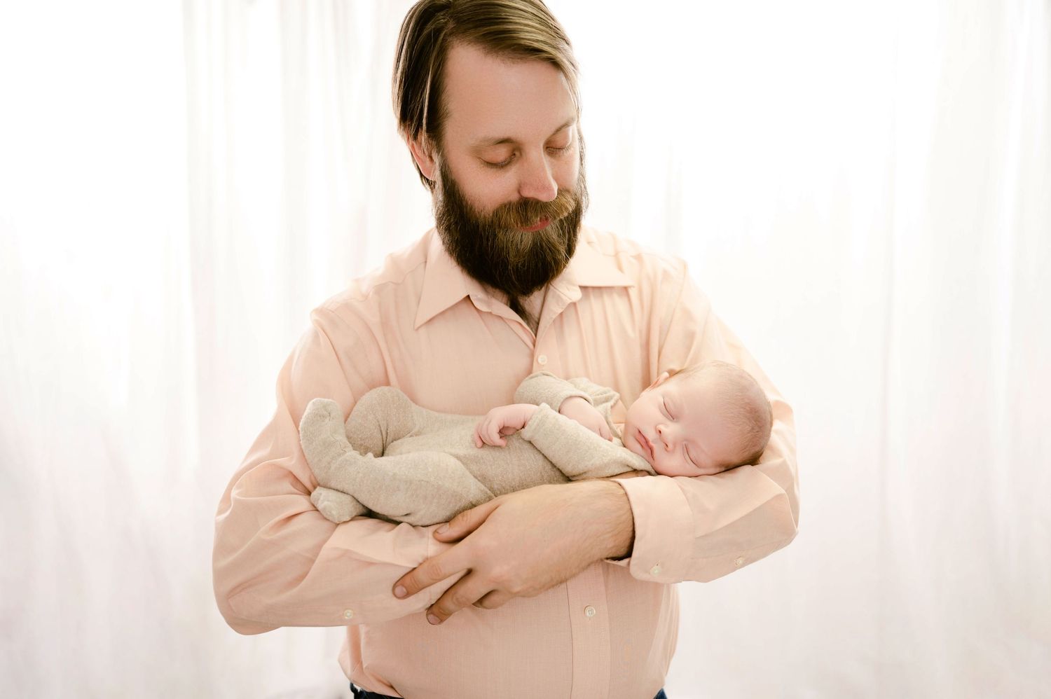 A person in a light colored shirt holds a sleeping newborn baby against their chest in front of a white backdrop.