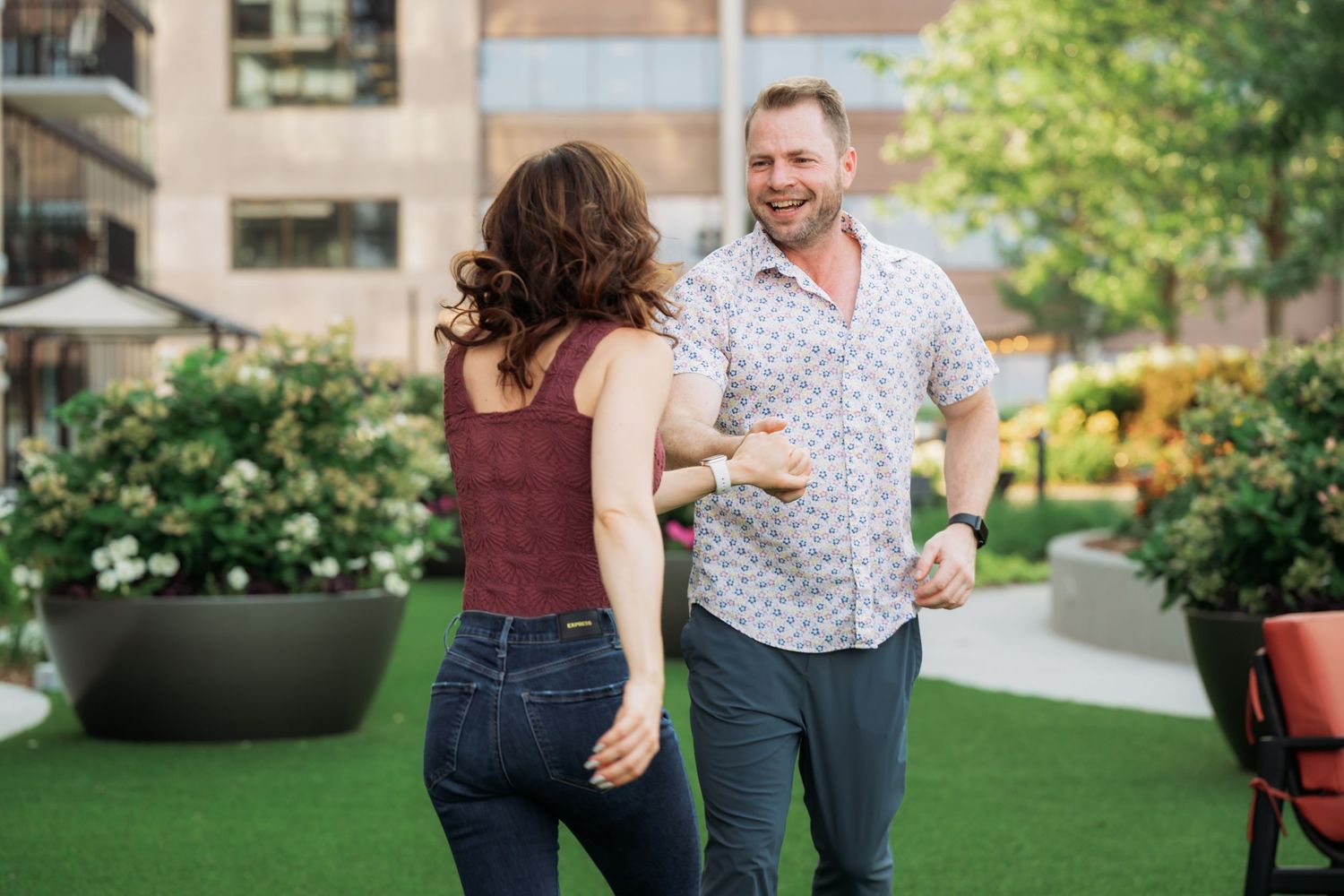 A couple dances and laughs together on a green lawn with buildings in the background.
