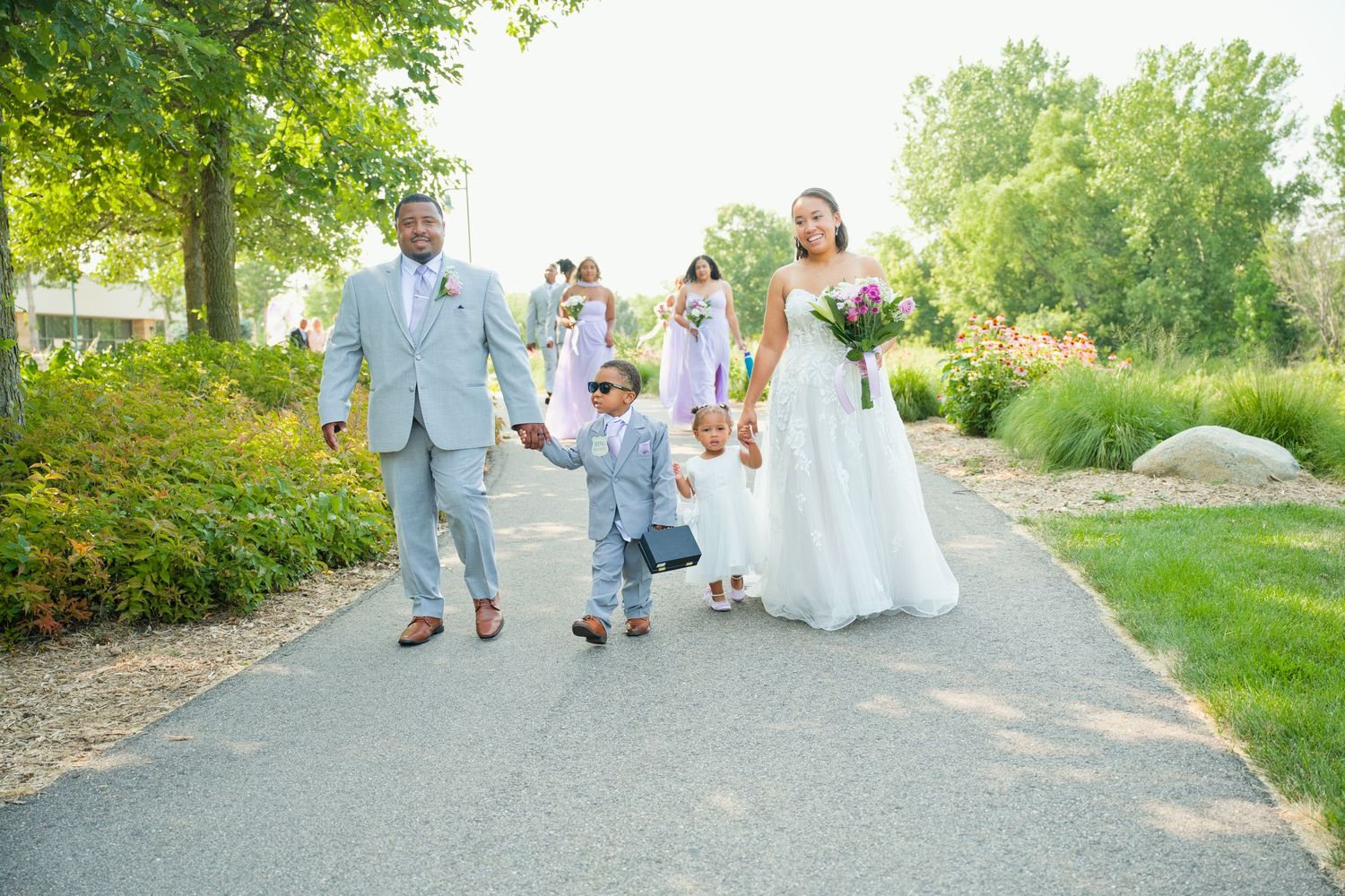 Bride and groom walk path with young boy in gray suit at outdoor wedding.
