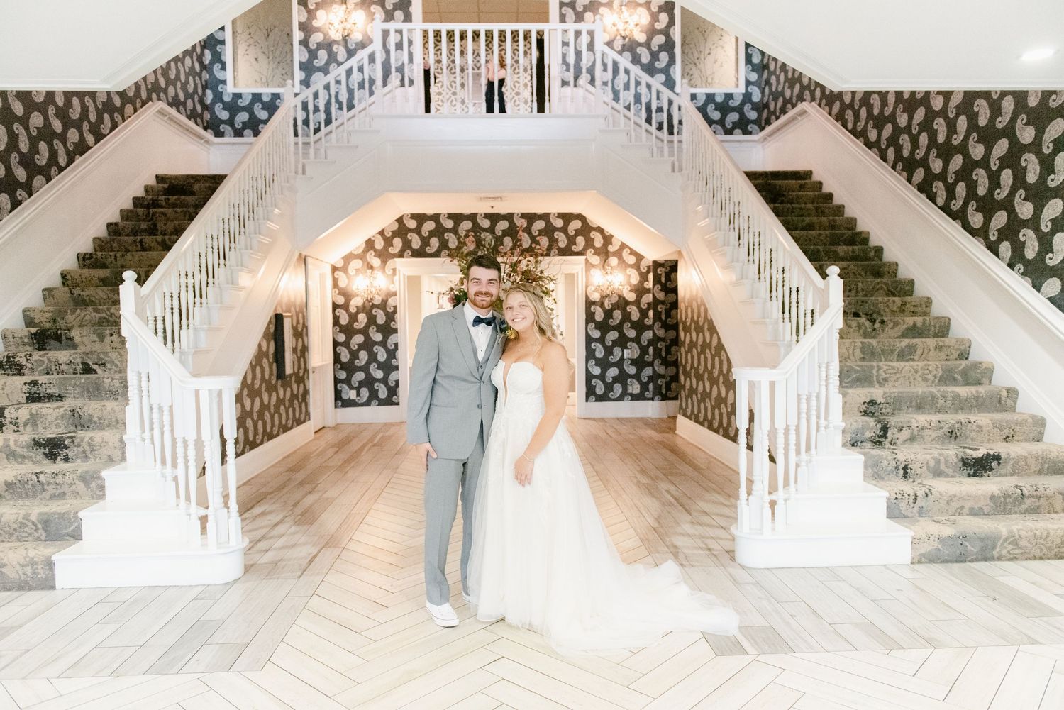 Central MN Wedding Photos | Bride and groom pose together at base of grand double staircase with string lights and bold wallpaper.