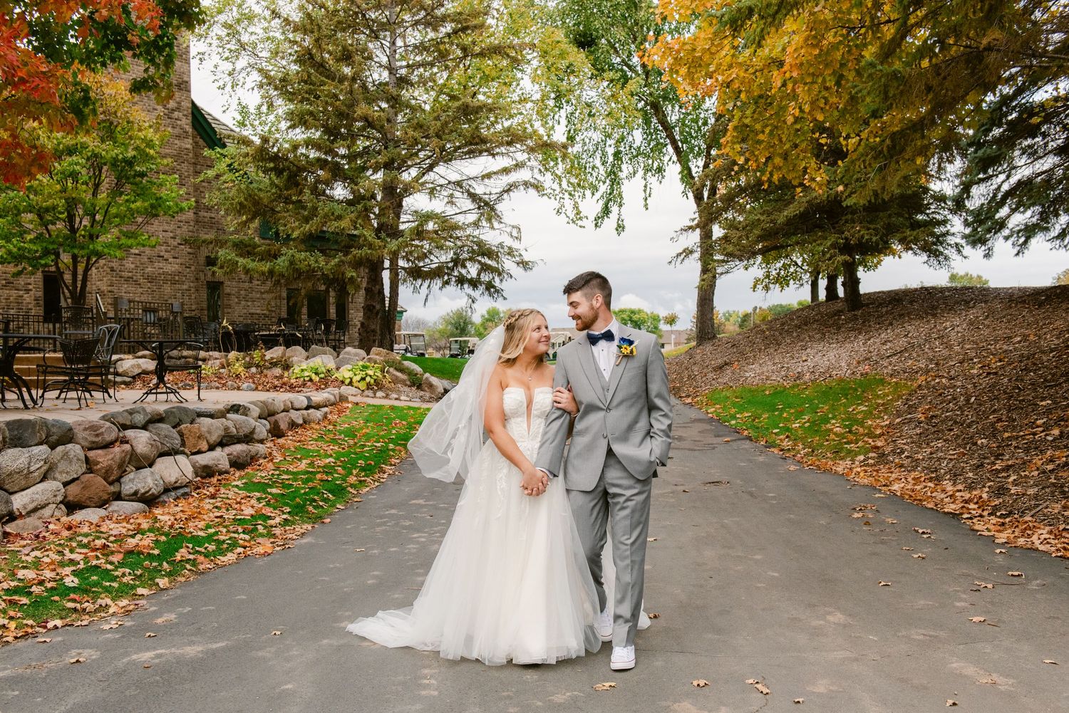 Central MN Wedding Photos | Bride and groom walking hand in hand on autumn tree-lined wedding venue path.