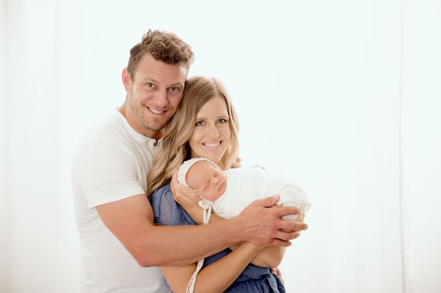Happy couple smiling and holding their newborn baby, posing together against a bright white background.