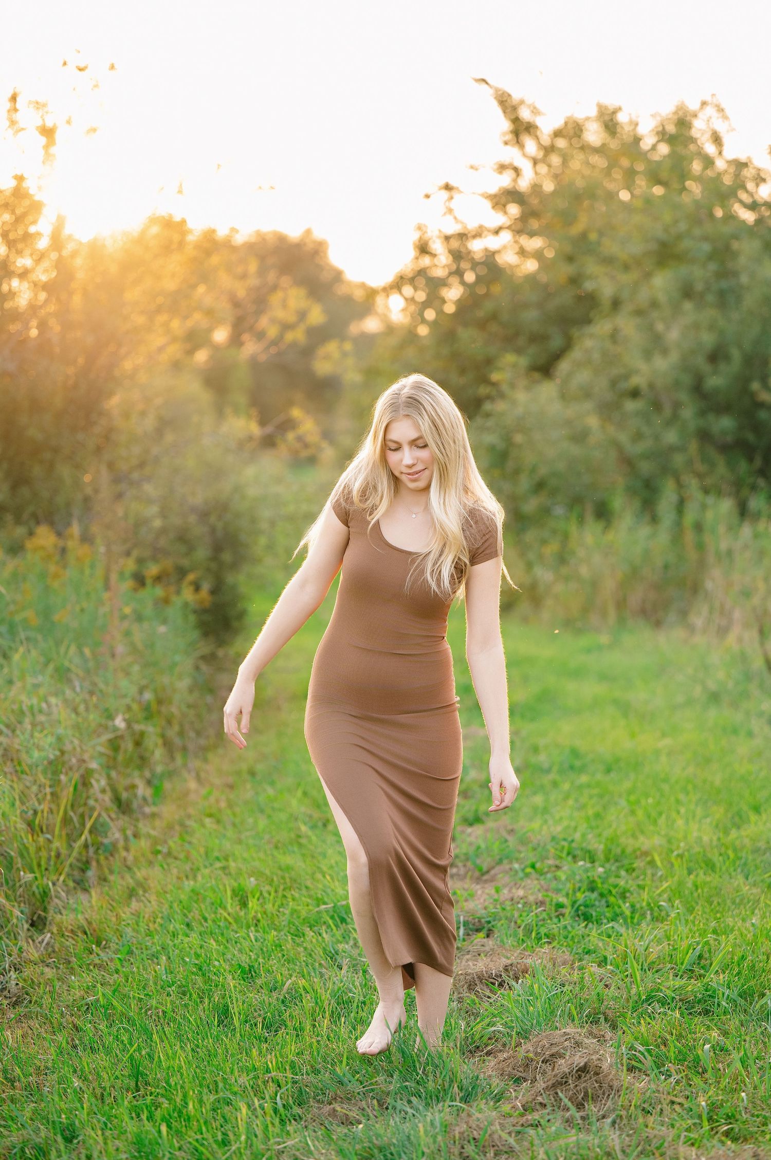 Central MN Senior Photography - Blonde woman in brown midi dress walking barefoot through lush green field at golden hour sunset.