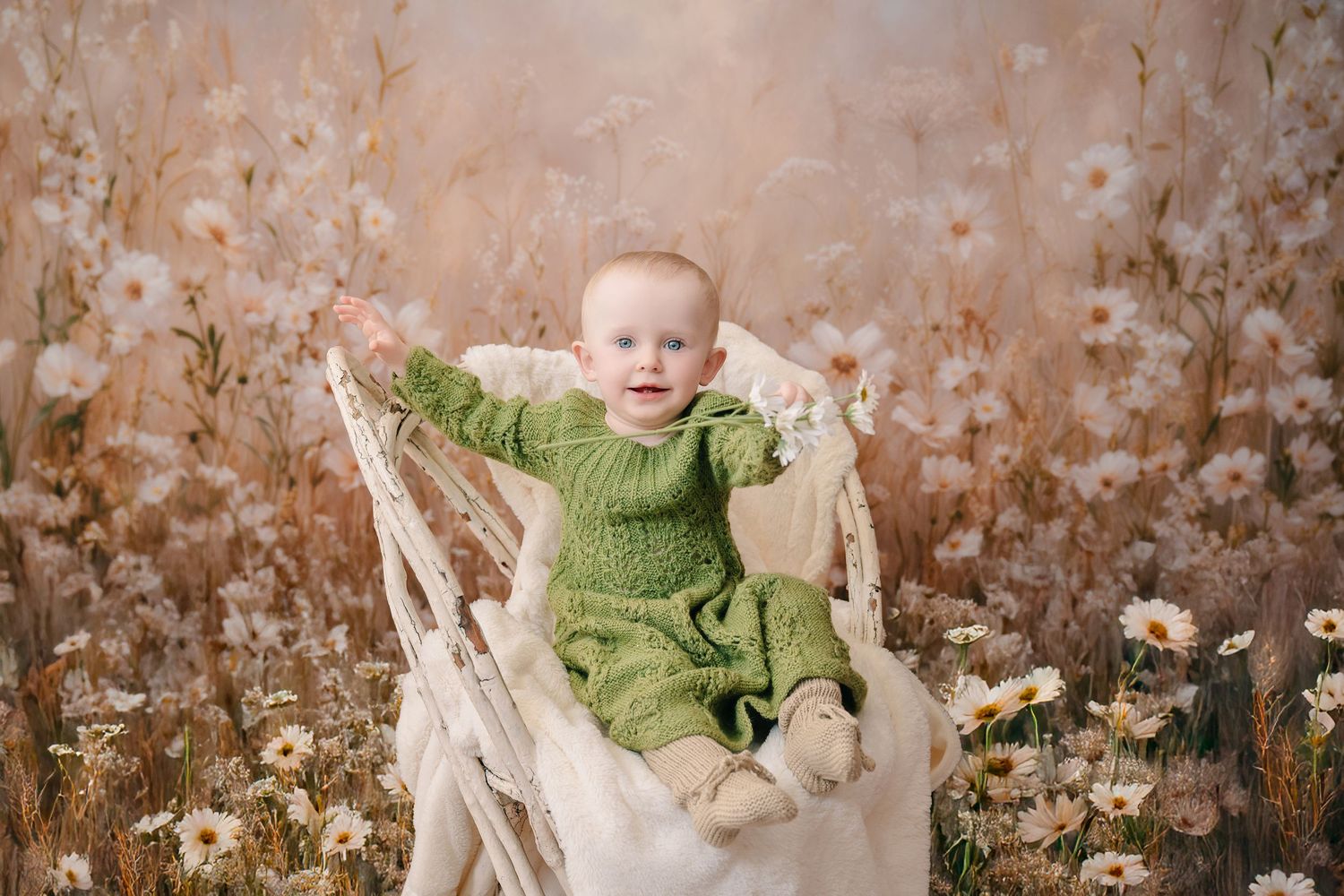 Adorable baby in green knit romper sitting in white chair surrounded by daisy wildflower field backdrop. mother day photoshoot