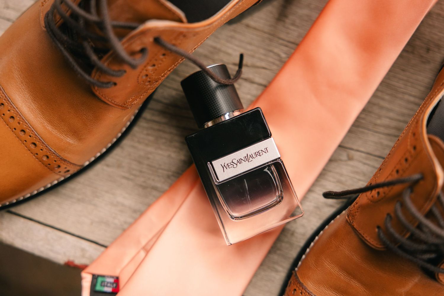 YSL cologne bottle beside tan leather brogue shoes and an orange tie on a wooden surface.