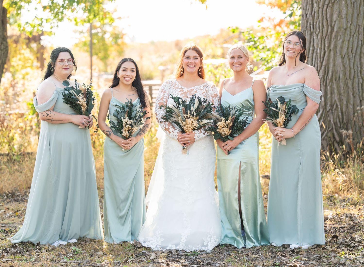 Bride with four bridesmaids in sage green gowns holding dried floral bouquets in autumn setting.