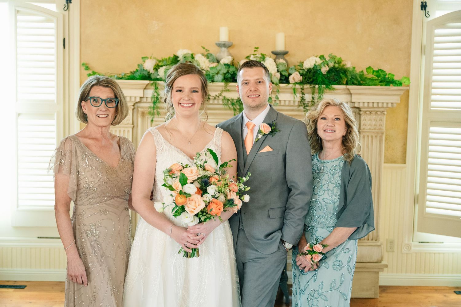 Wedding portrait of bride and groom with two mothers in formal attire in front of a floral fireplace.
