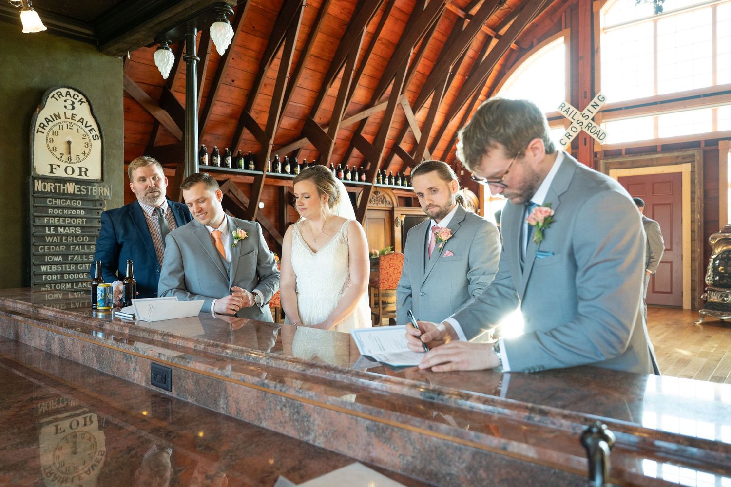 Bride and groom signing marriage documents at wooden bar counter with witnesses beside them.