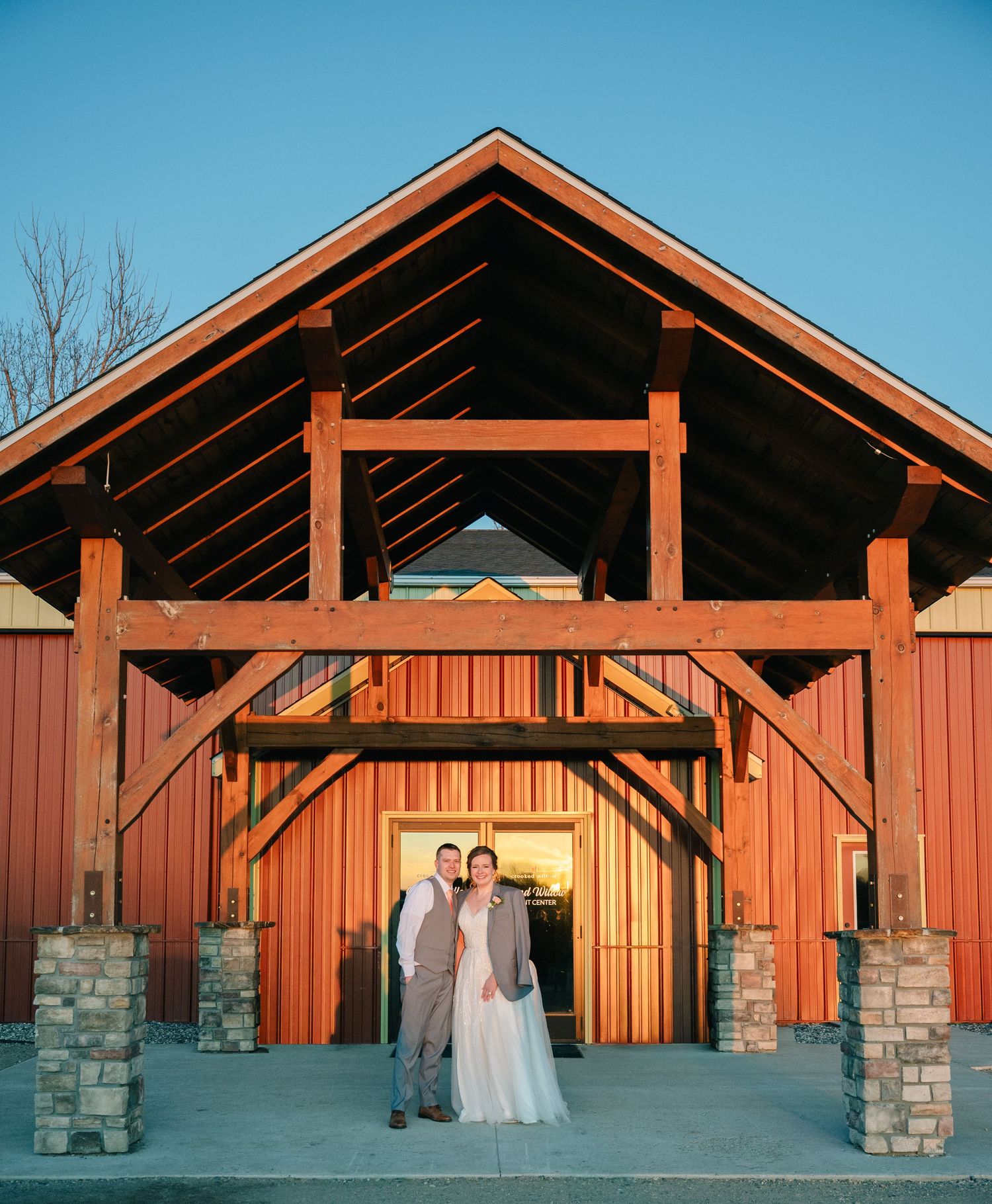 Bride and groom pose together at entrance of rustic red barn wedding venue at dusk.