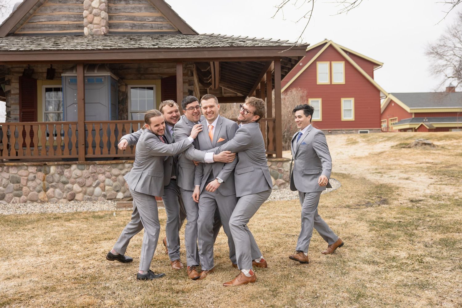 Groomsmen in gray suits playfully huddle around groom outside rustic barn wedding venue.