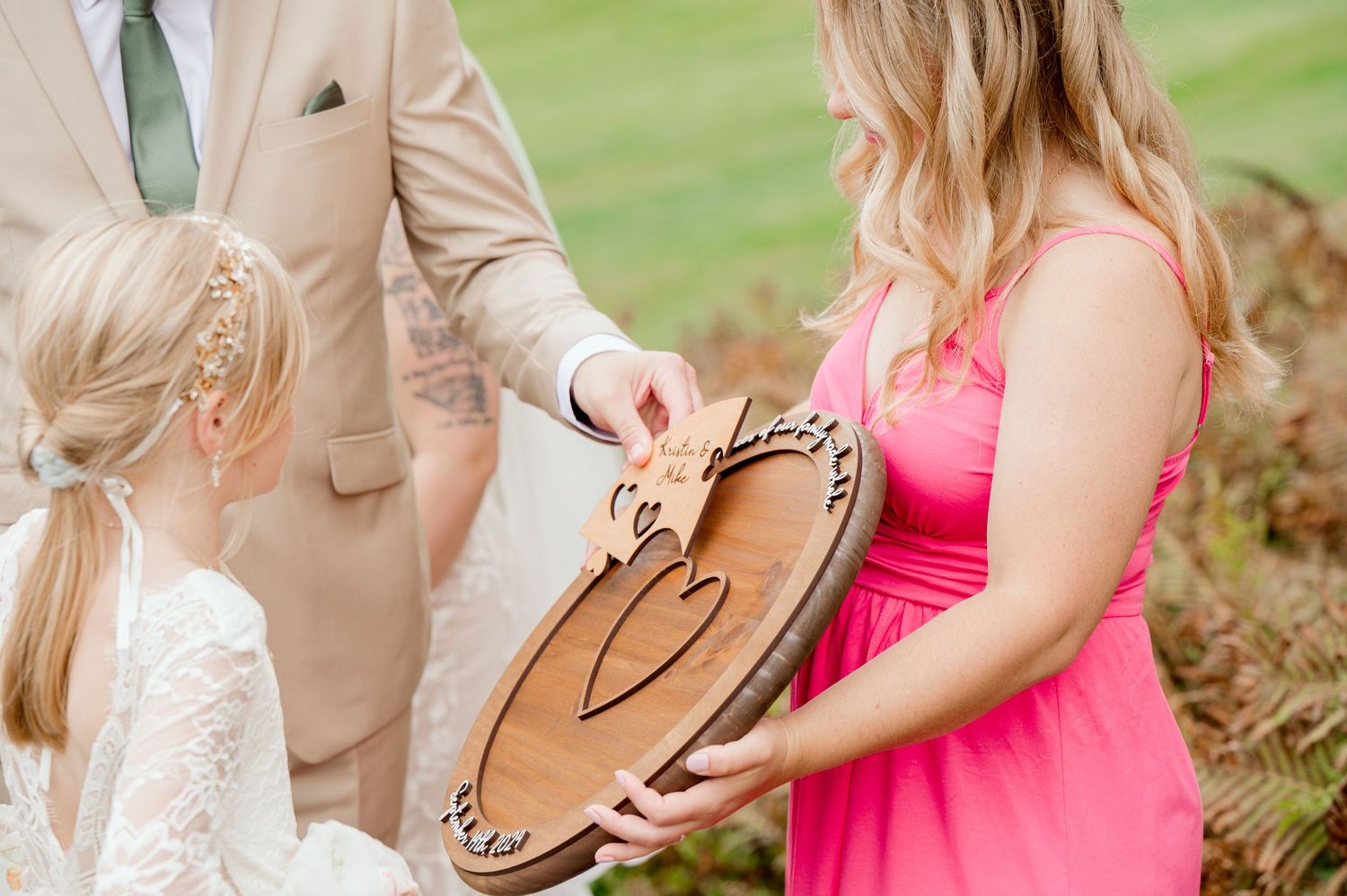 Wedding party members examine a decorative wooden ring holder during outdoor ceremony.