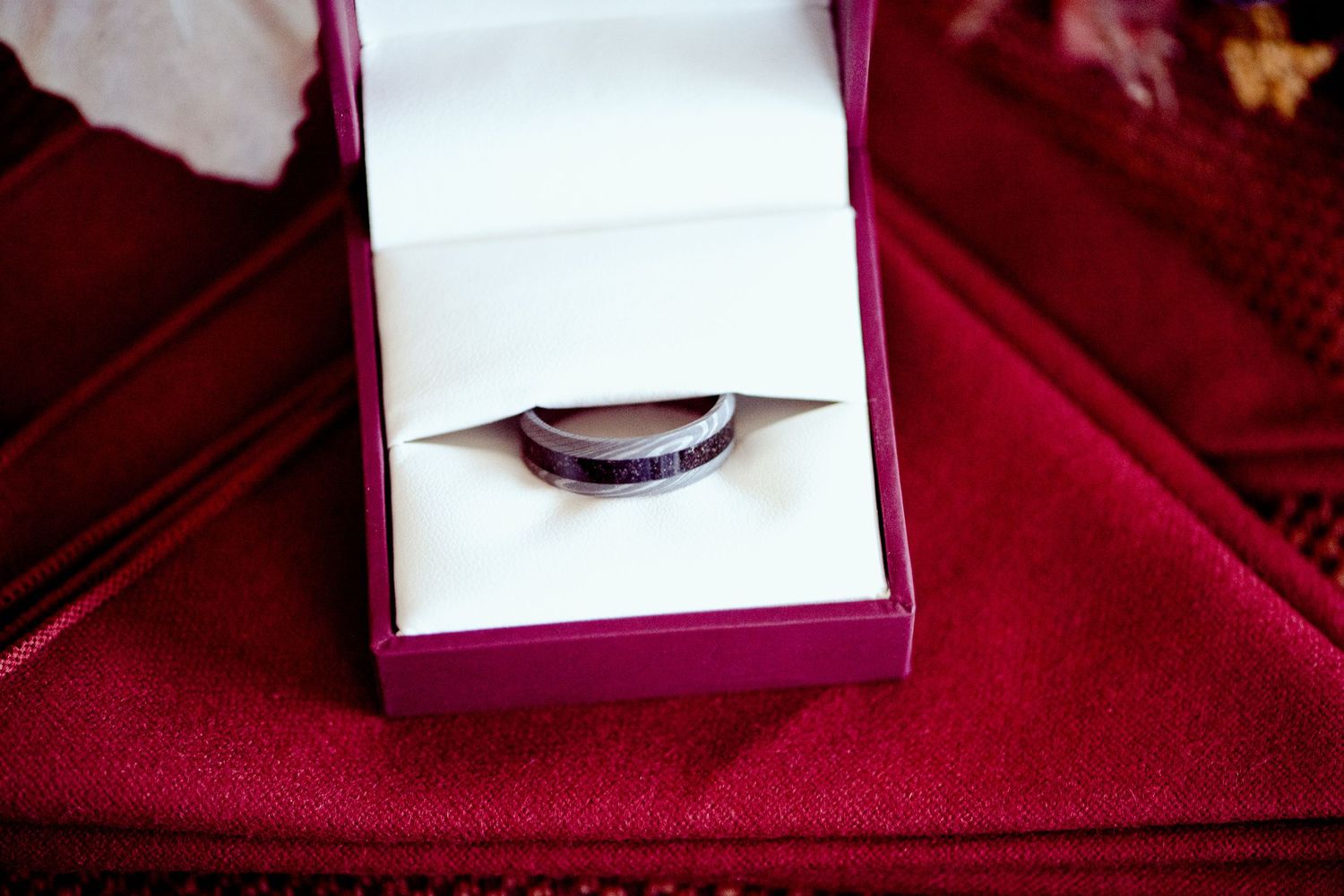 A silver wedding ring nestled in a burgundy velvet box photographed against red fabric.
