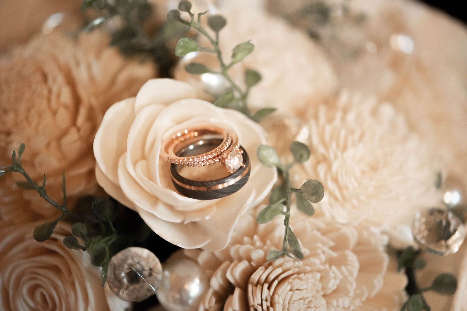 Two gold wedding rings nestled among delicate cream roses and eucalyptus greenery arranged in a romantic bridal display. Princeton MN Milest