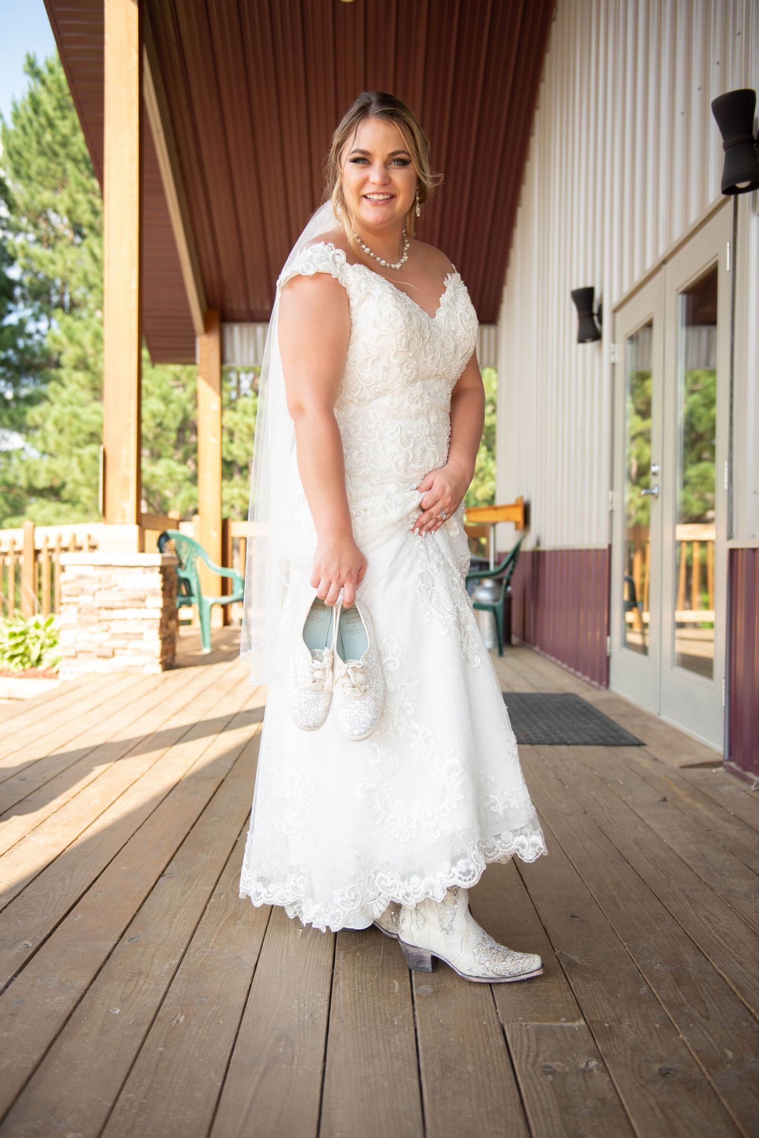 A bride in a white lace gown and cowboy boots stands on a rustic wooden porch. Zimmerman MN Weddings