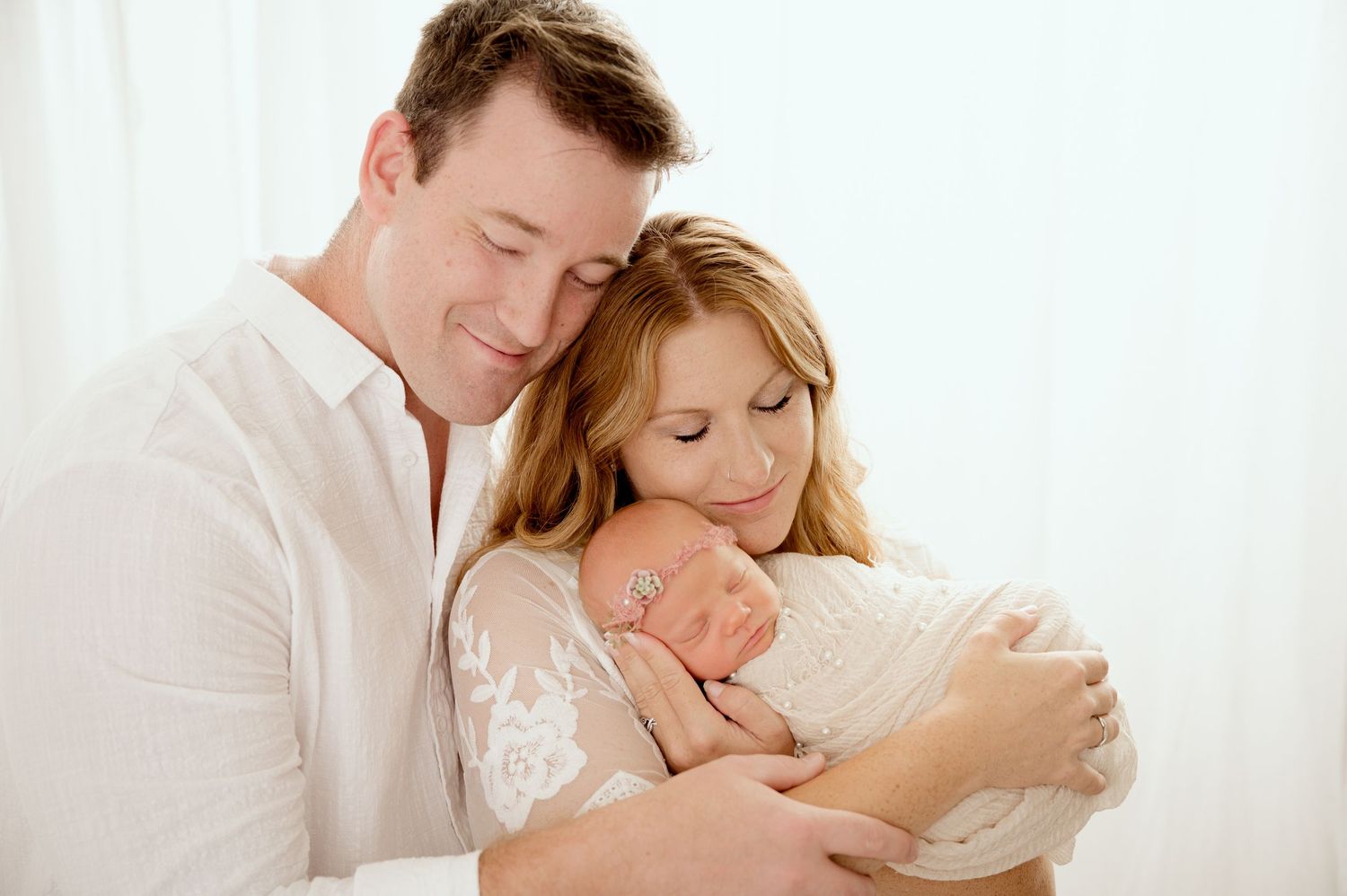 New parents in white outfits tenderly holding their newborn baby during a professional photo session.