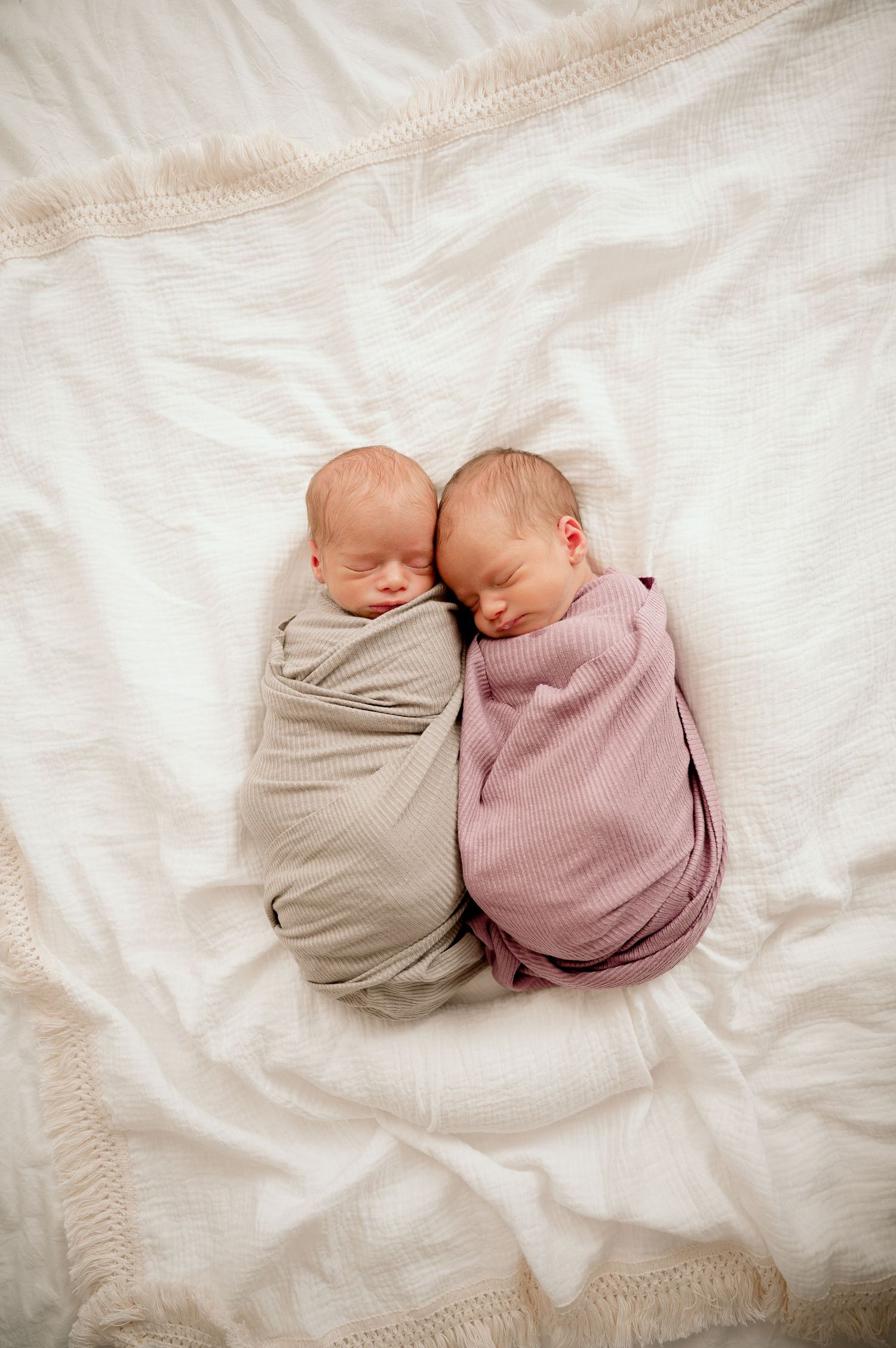 Two swaddled newborn twins sleeping peacefully side by side on a white blanket, wrapped in gray and pink muslin cloths.