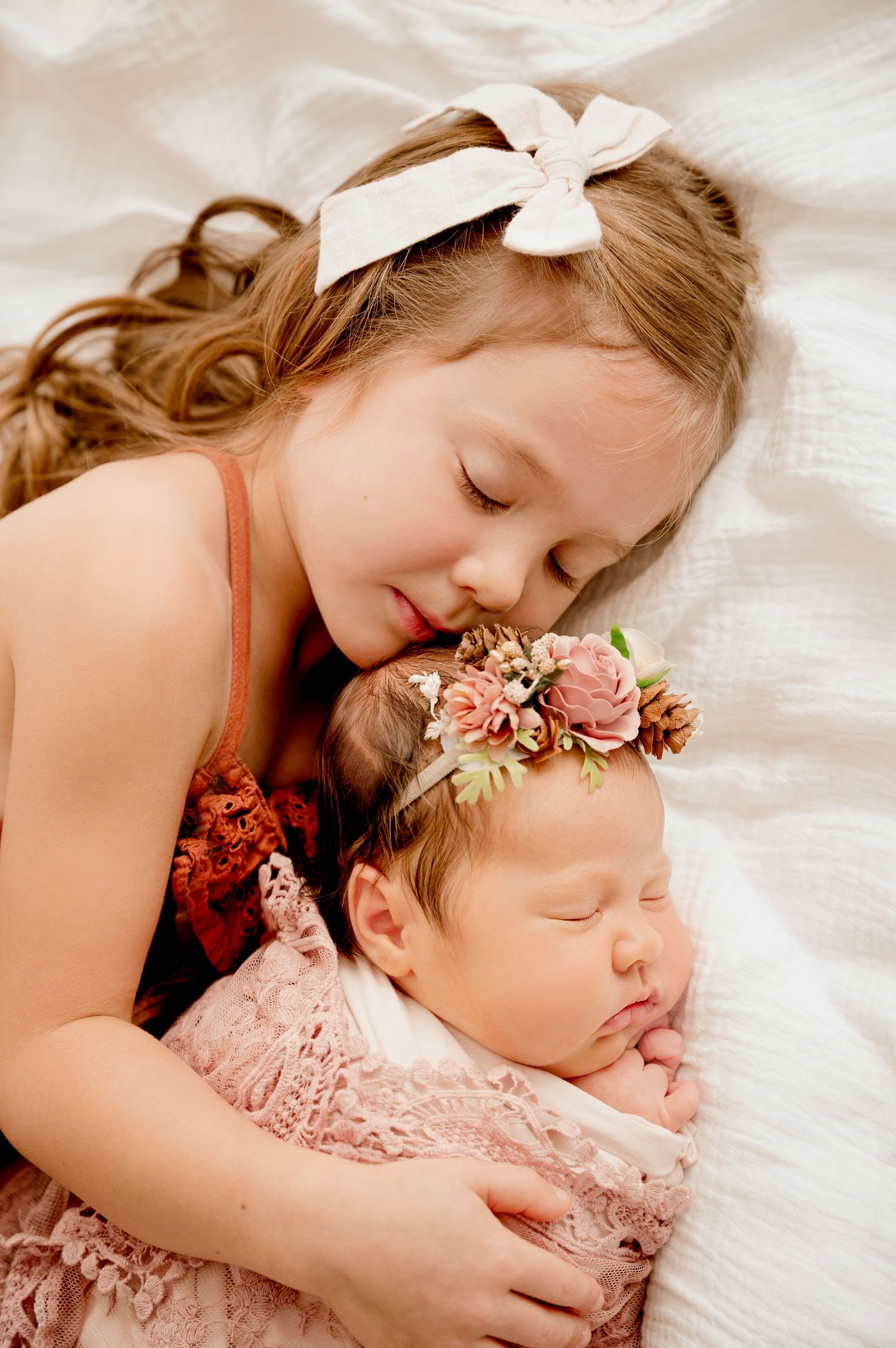 Older sister with white bow gently holds sleeping newborn baby girl wearing floral headband in pink lace wrap.