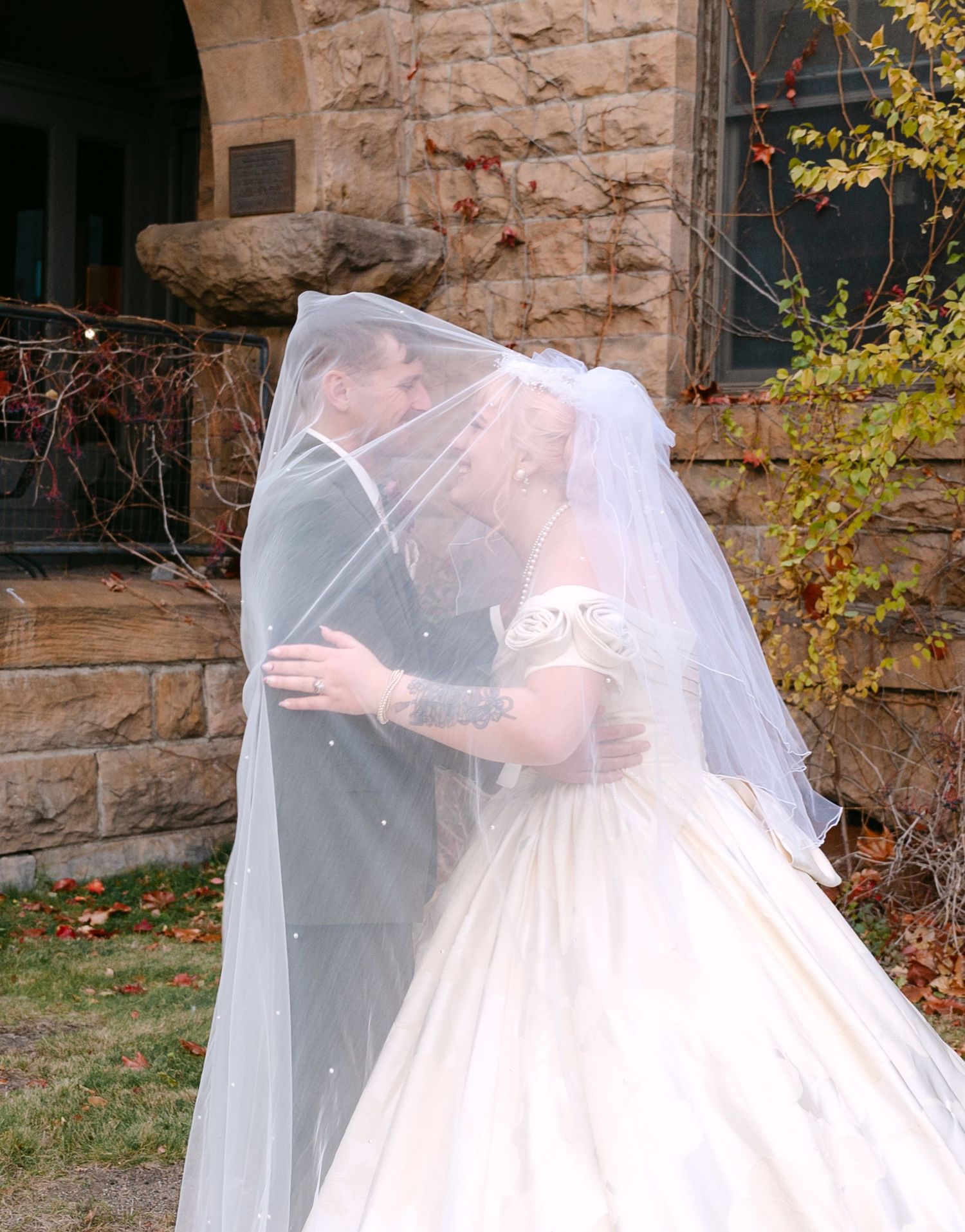 Central MN Wedding Photos | Bride and groom share romantic kiss under flowing white veil outside historic stone building.