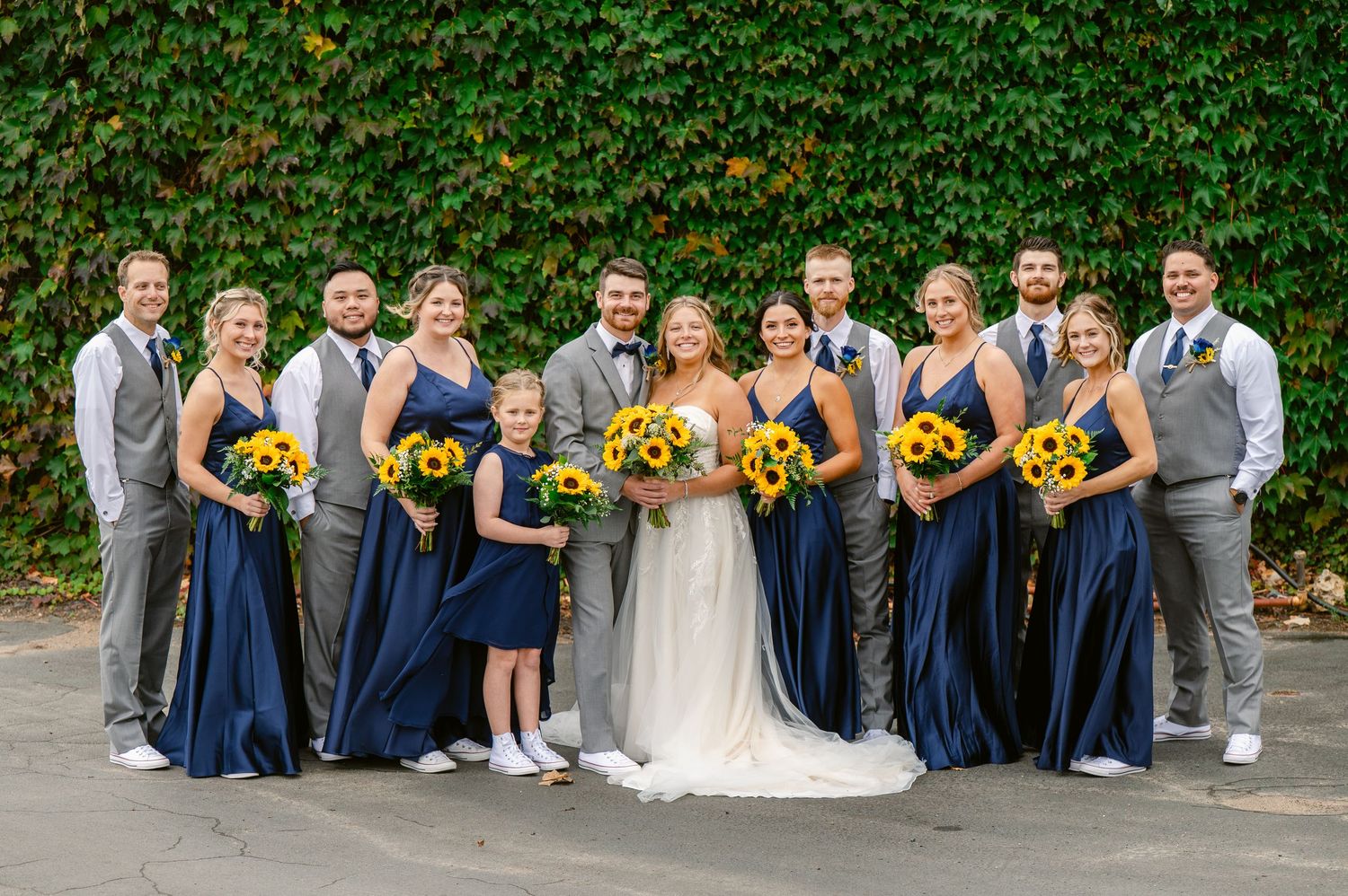 Central MN Wedding Photos | Wedding party poses in navy and gray attire with sunflower bouquets in front of a green ivy-covered wall.