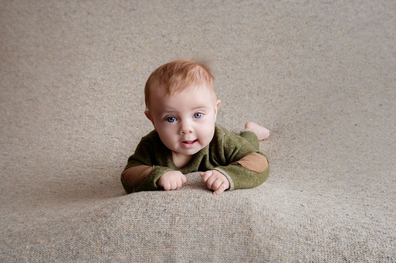 Central MN Milestone PhotographyAdorable red-haired baby in green onesie doing tummy time on beige carpet, looking up with bright blue eyes.