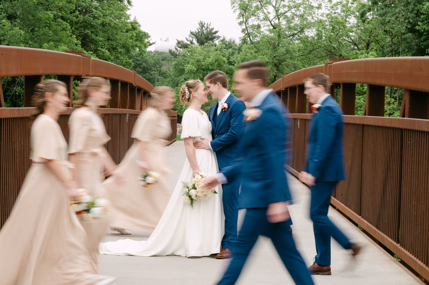 Wedding party in blue and blush attire walks across wooden bridge in outdoor garden setting.