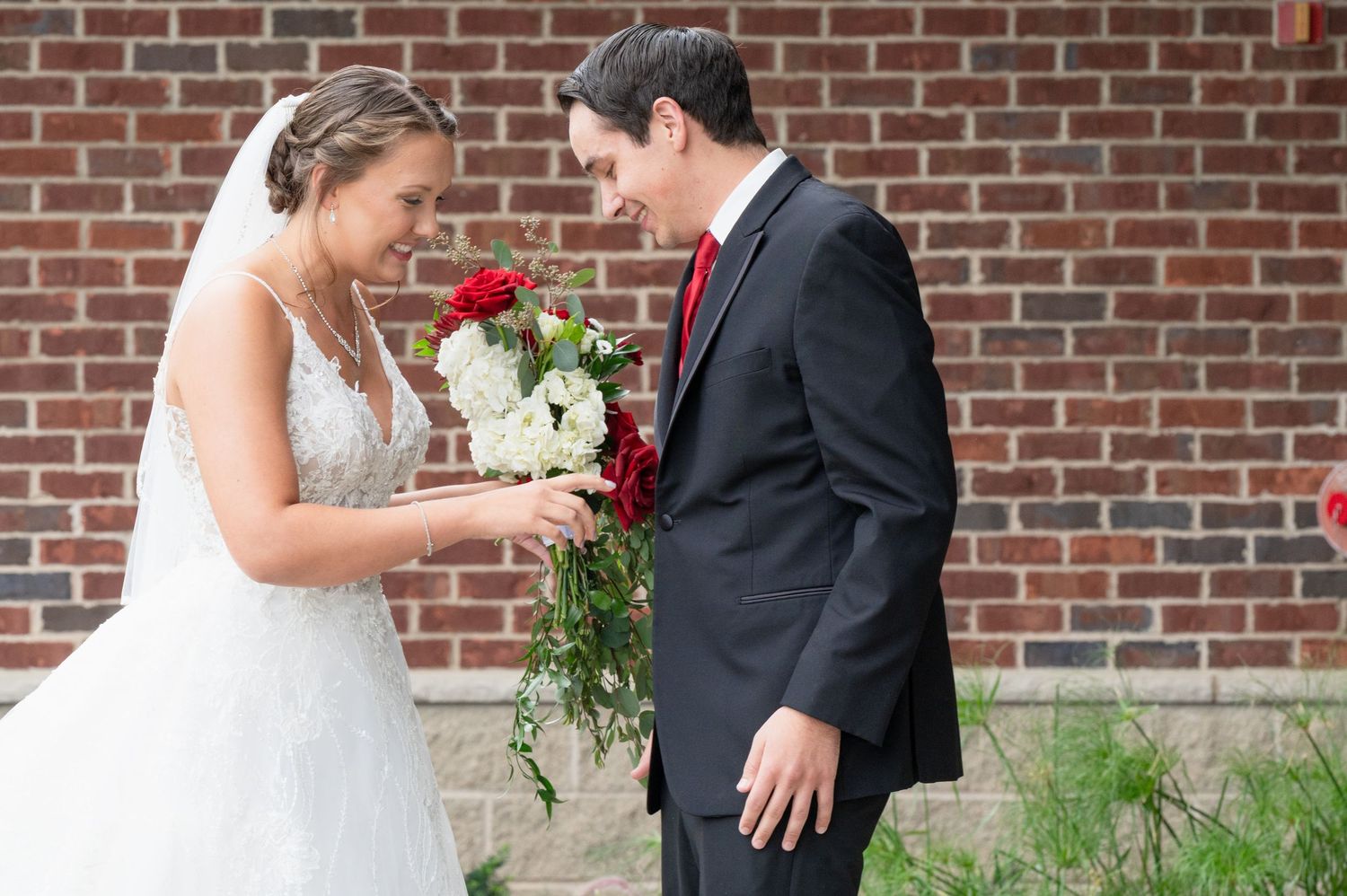 Bride and groom share a tender moment examining wedding bouquet of red and white roses outside brick building.