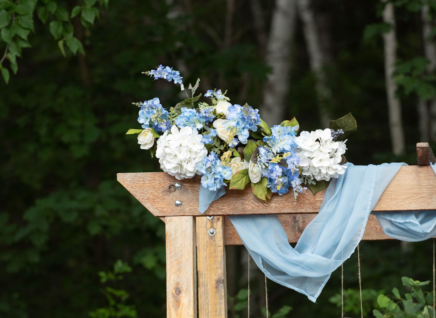 A rustic wooden archway adorned with light blue fabric and delicate flowers creates an outdoor wedding ceremony setting. Zimmerman MN Weddin