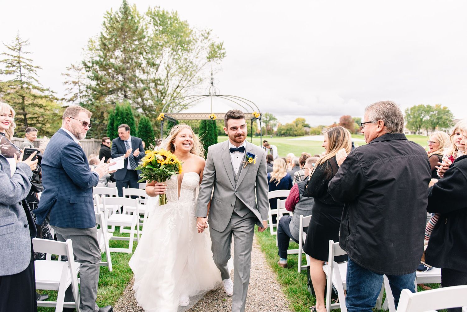 Central MN Wedding Photos - Joyful newlyweds walk back down aisle after ceremony, bride holding sunflower bouquet, guests applauding.