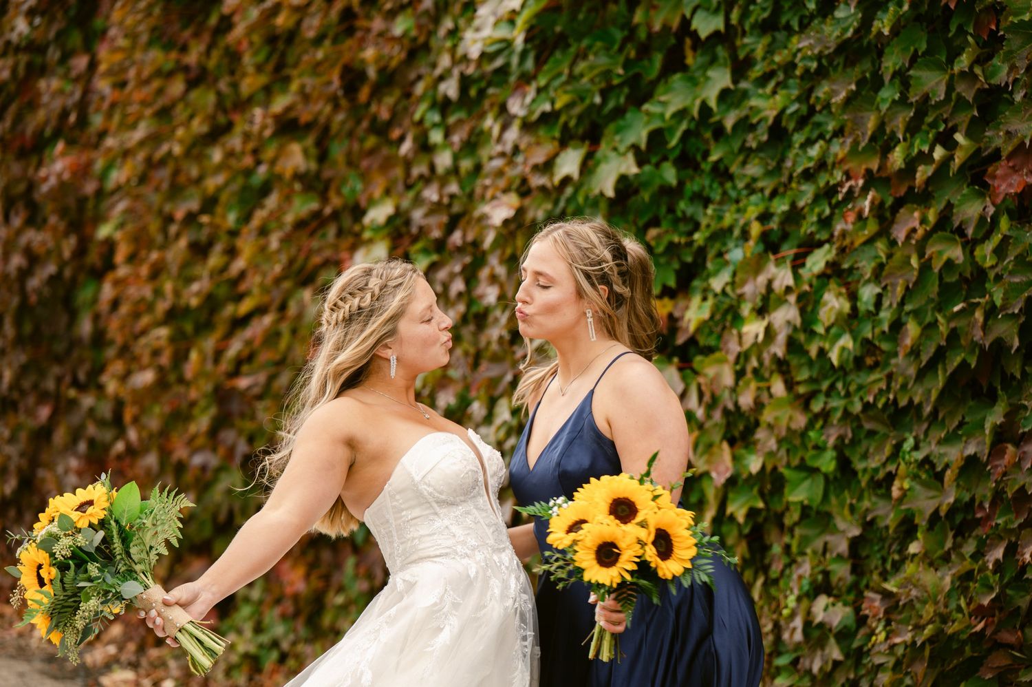 Central MN Wedding Photos | Bride and bridesmaid holding sunflower bouquets in front of ivy wall.