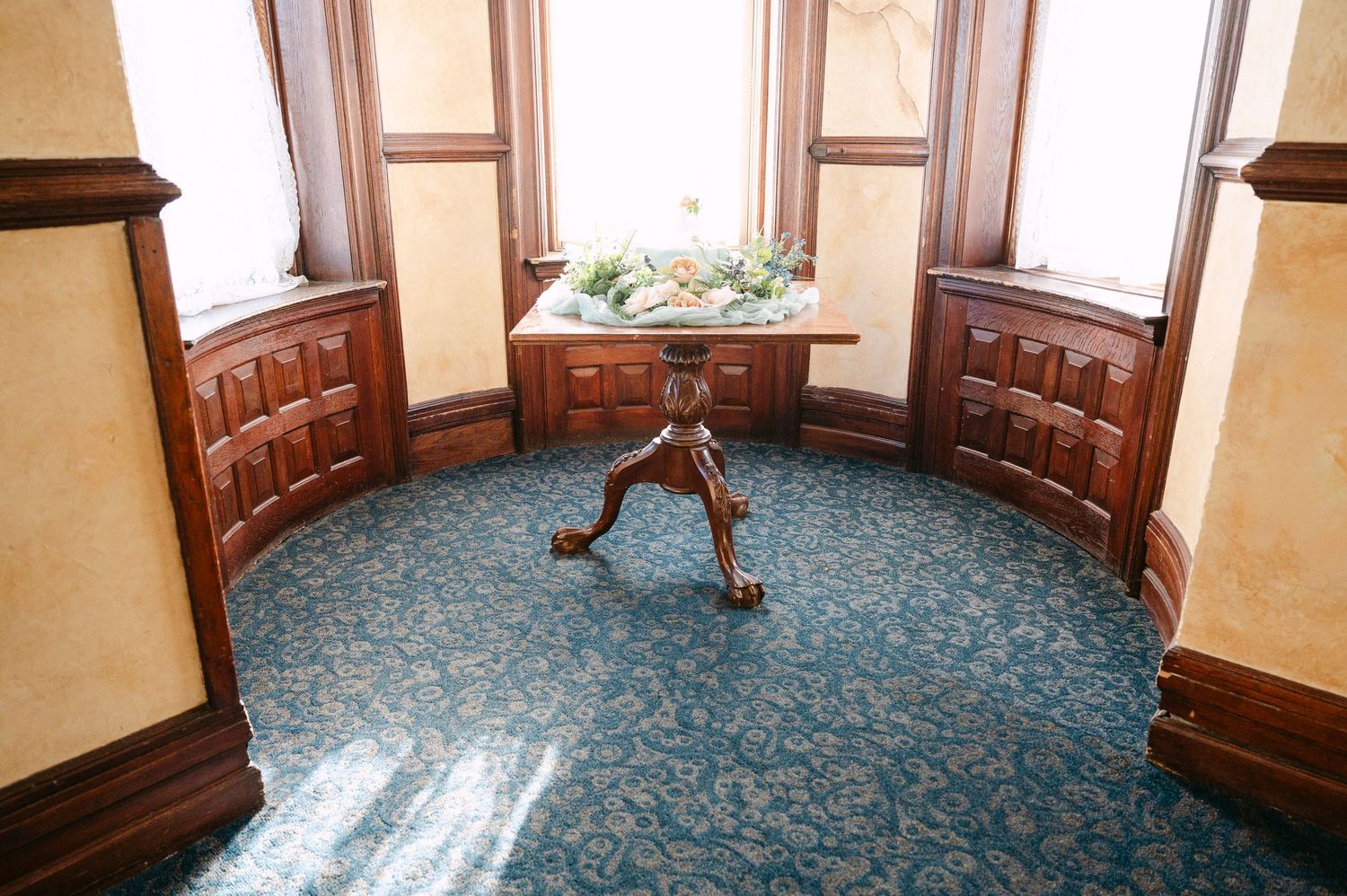 Central MN Wedding Elegant bay window nook with ornate wooden trim, blue floral carpet, and antique pedestal table with floral arrangement.