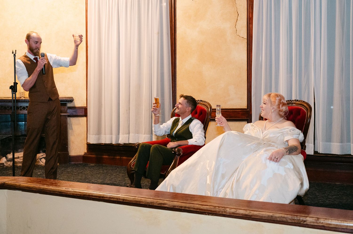 Central MN Wedding Photos | Best man gives toast while bride and groom relax in velvet chairs during wedding reception speech.
