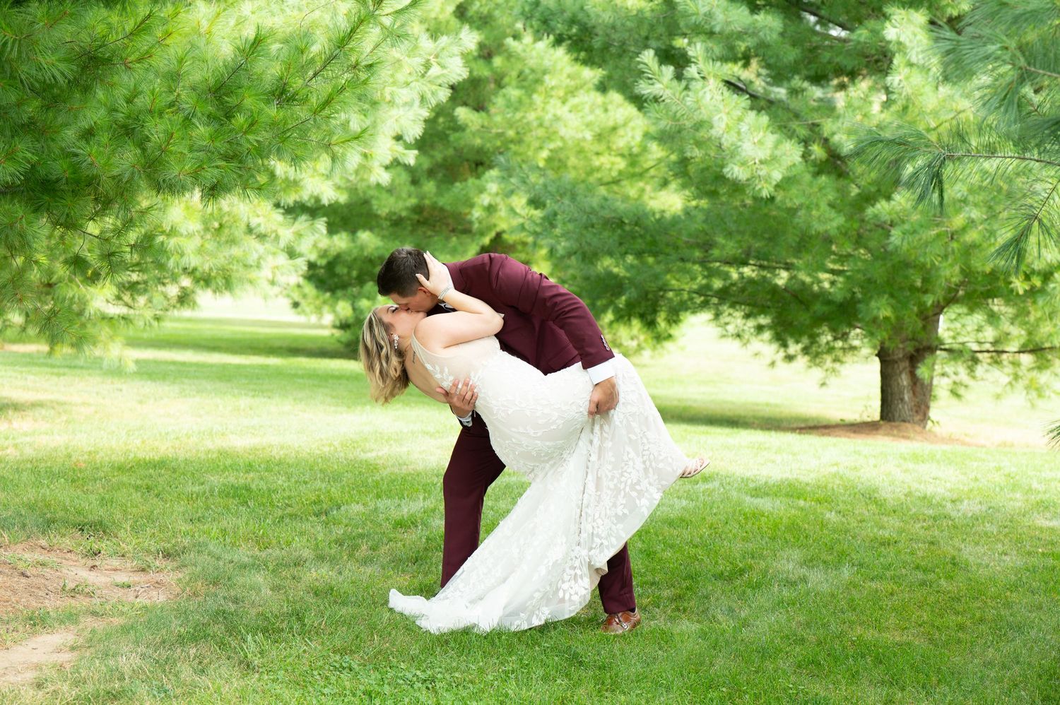 Central MN Wedding Photography - Groom in burgundy suit dips bride in white lace wedding dress for a romantic kiss outdoors.