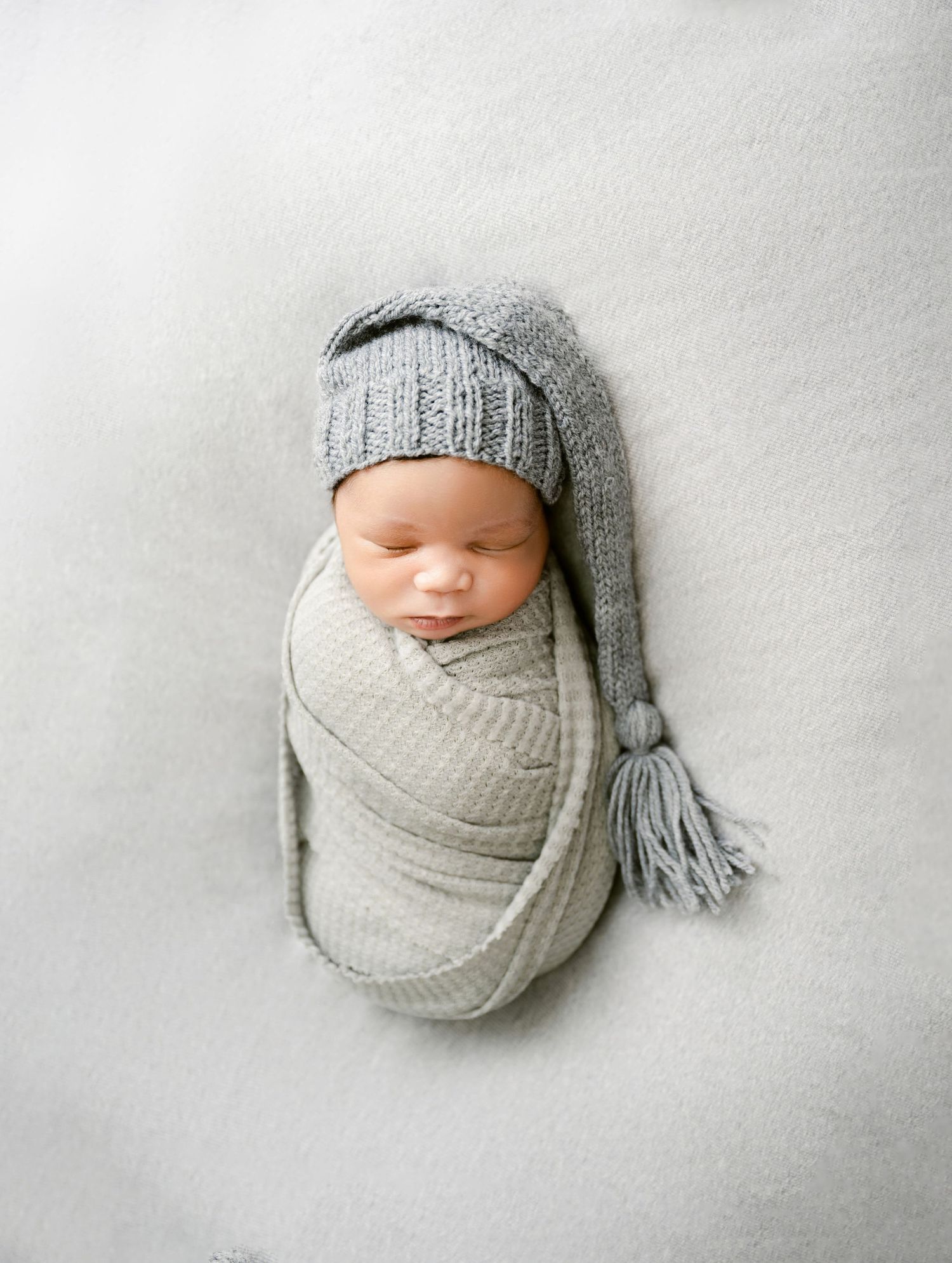 Sleeping newborn swaddled in gray wrap wearing a knit tassel hat on a soft white background.