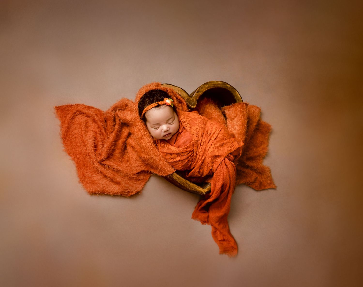 Sleeping newborn wrapped in orange cloth inside a heart-shaped wooden bowl prop.