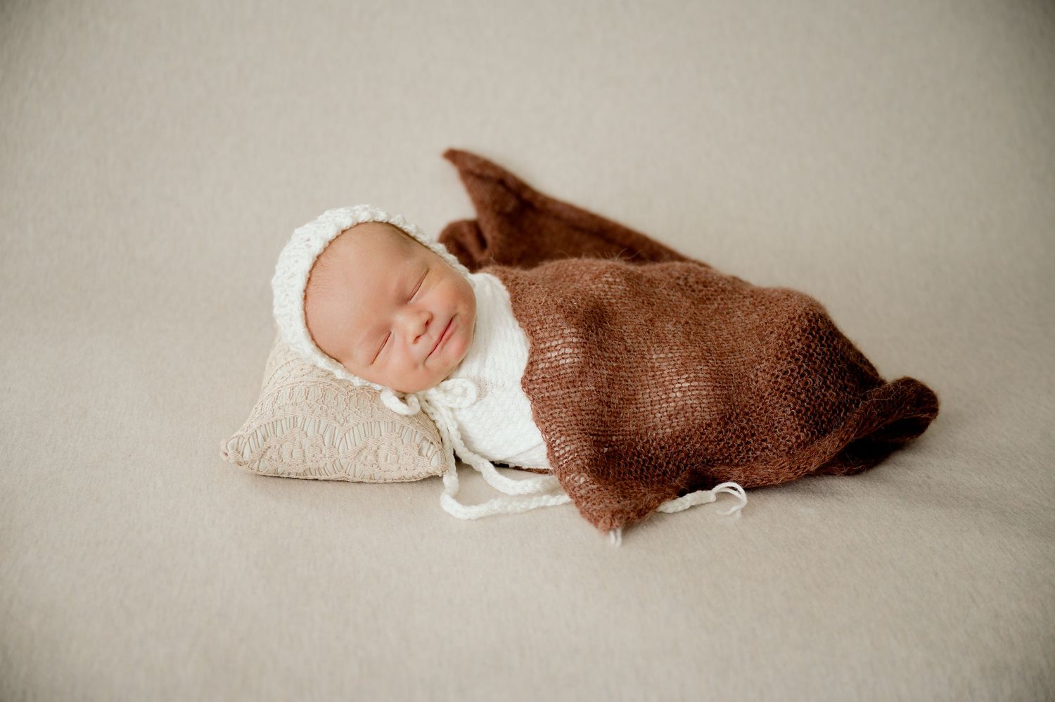 Sleeping newborn baby wrapped in brown knit blanket wearing white bonnet on beige backdrop.