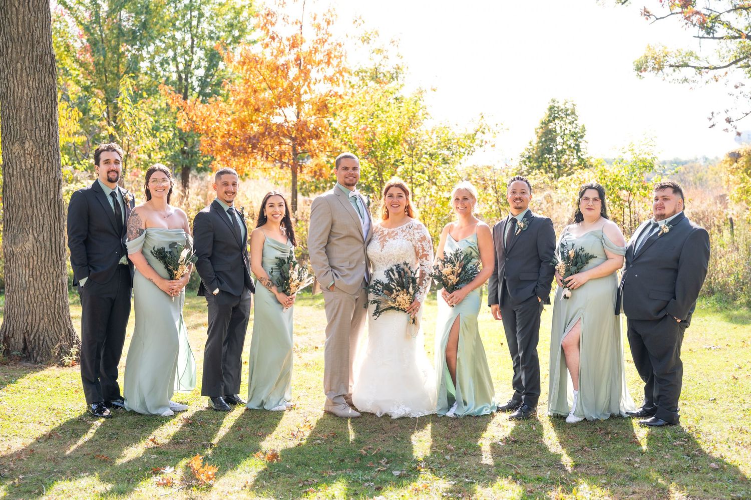 Wedding party of nine poses on a sunny fall day, bridesmaids in mint green dresses and groomsmen in dark suits.