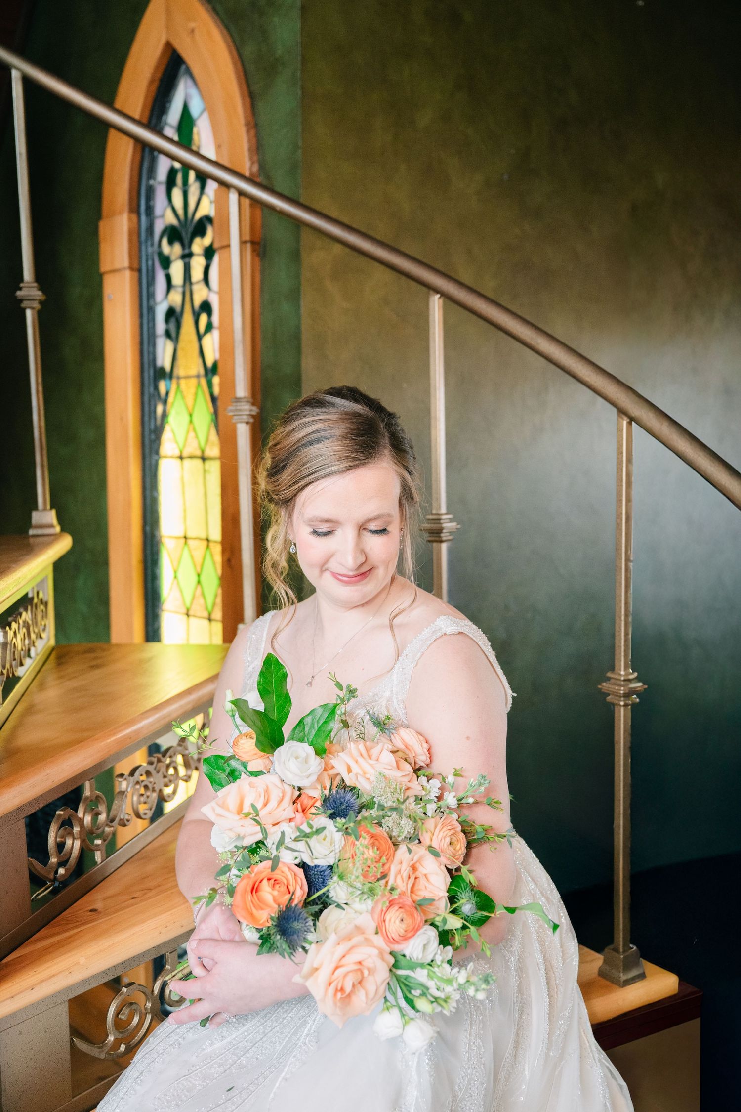 Bride in white gown holding a peach and blue floral bouquet on a staircase near stained glass.