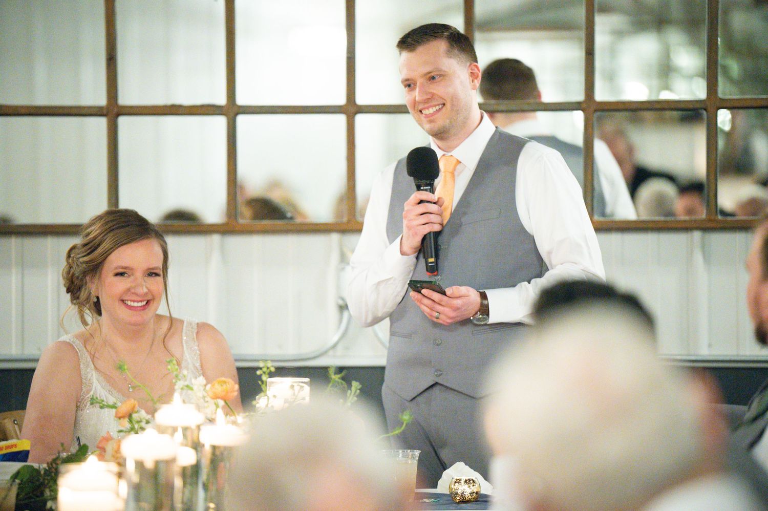 Smiling groom delivers wedding speech into microphone as bride laughs joyfully at elegant reception dinner.
