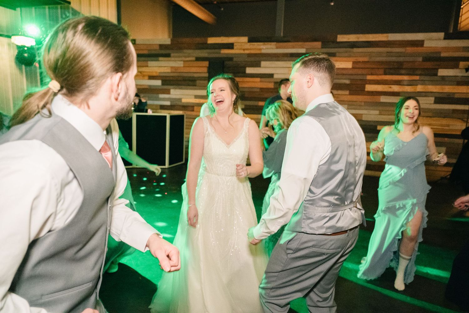 Bride in white gown laughs and dances with groomsmen under green lights at wedding reception.