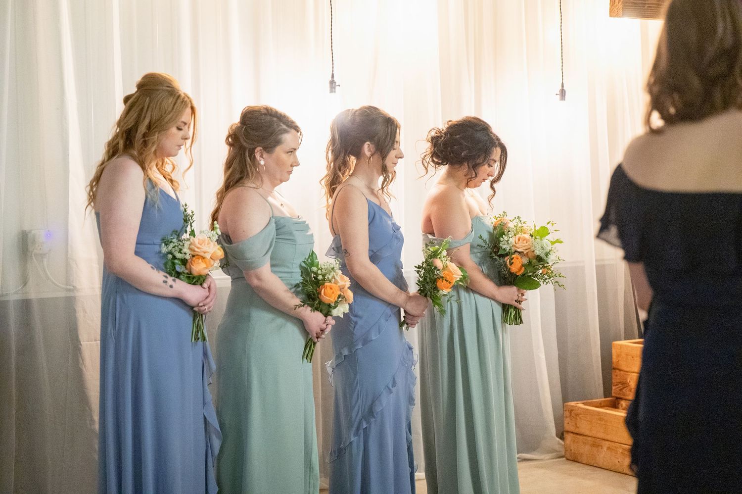 Four bridesmaids in blue and sage green gowns hold colorful bouquets during indoor wedding ceremony.