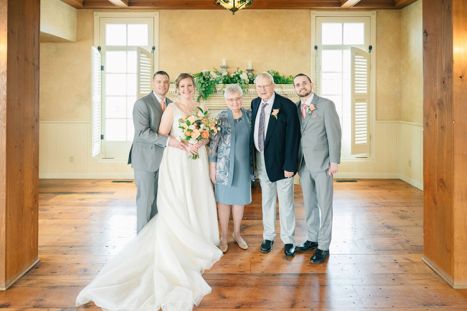 Bride with peach bouquet poses with four family members before floral fireplace display.