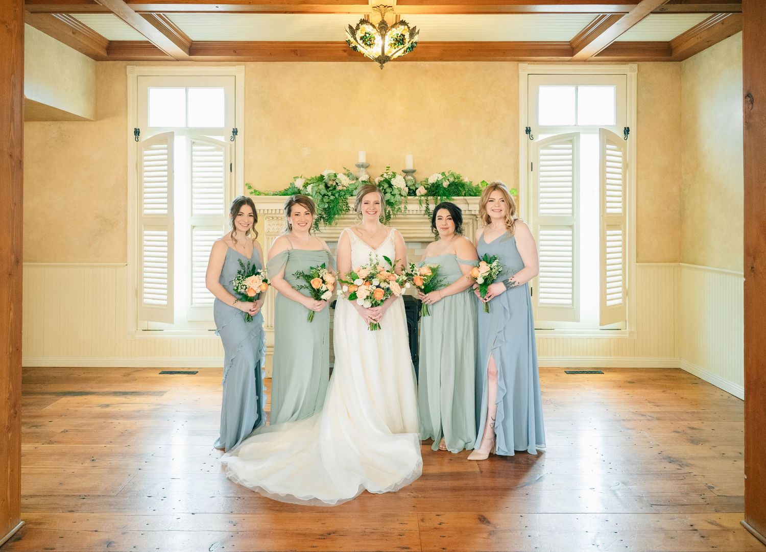 Bride in white gown with four bridesmaids in sage green dresses posing in elegant wood-floored room.