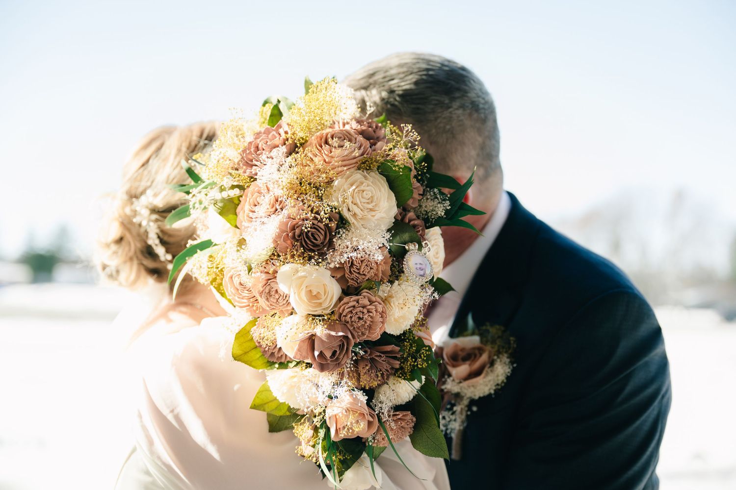 A wedding couple shares a tender moment outdoors with a soft pink and white floral bouquet in view.