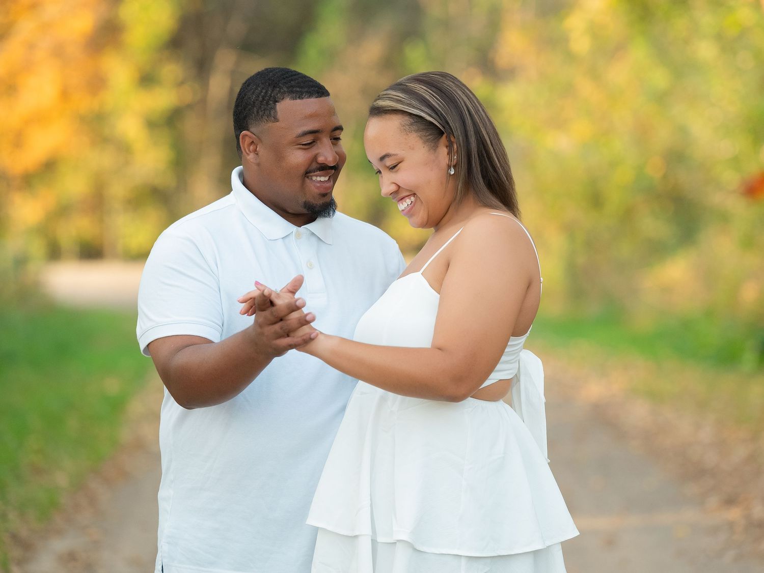 A couple wearing white outfits share a joyful moment together on a path with autumn foliage in the background.