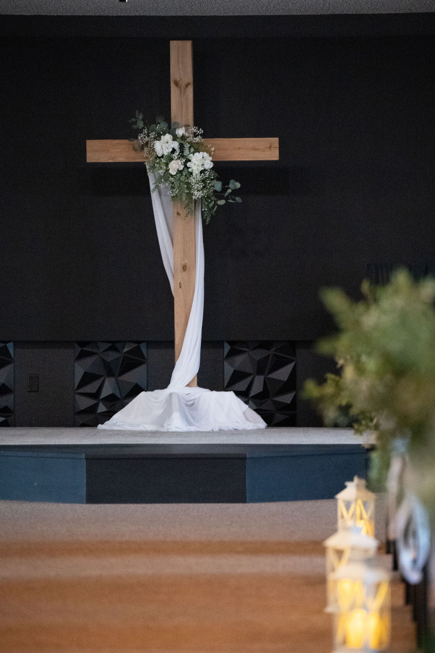 Wooden cross decorated with white fabric and floral arrangements against a dark background with a lantern in the foreground.