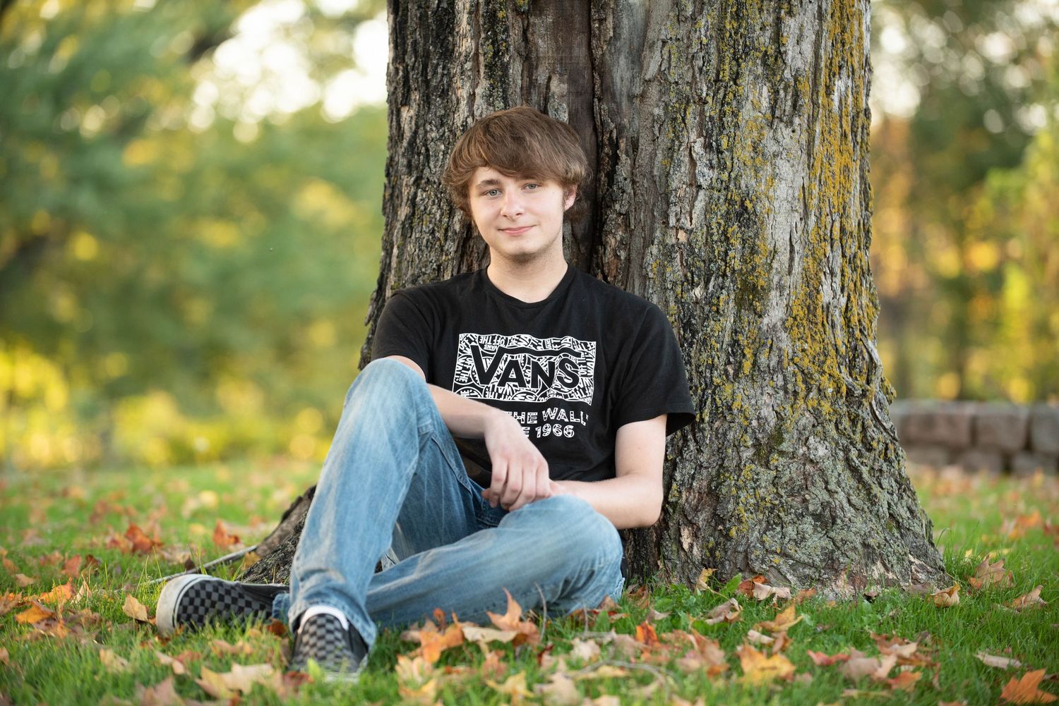 A person sits against a tree trunk in a sunlit forest setting wearing casual clothing.